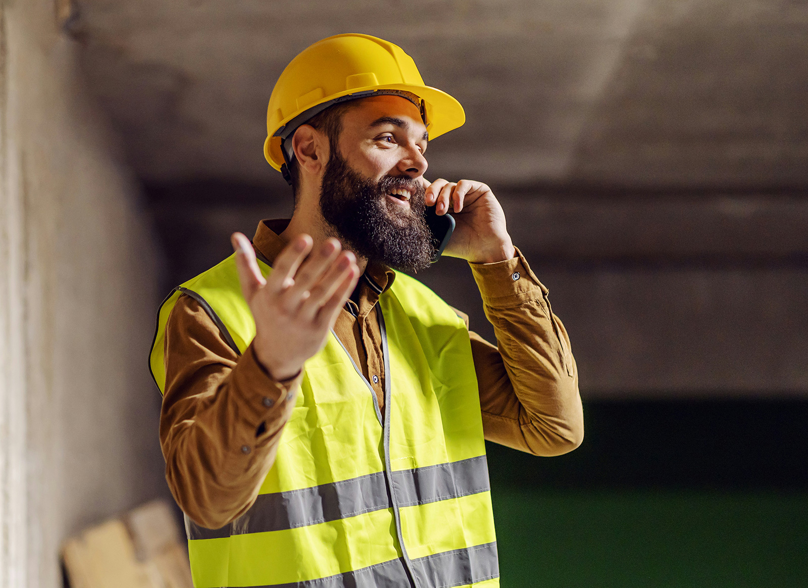 A contractor in a hard hat and high-vis vest taking a phone call on a job site, gesturing as he listens.